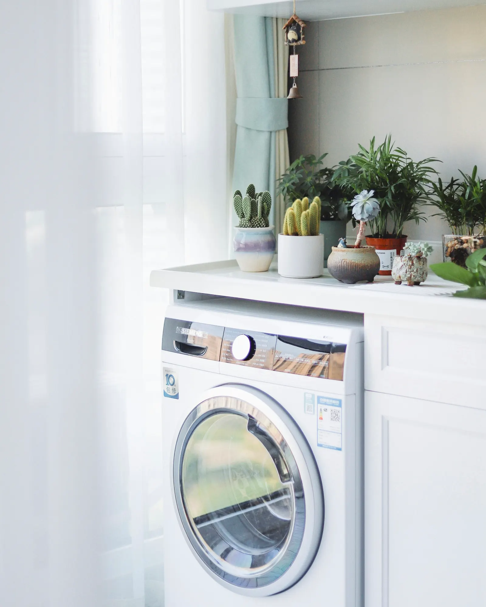 Front-loading washing machine built into a run of white cupboards on an apartment utility balcony, with a counter and potted plants above it