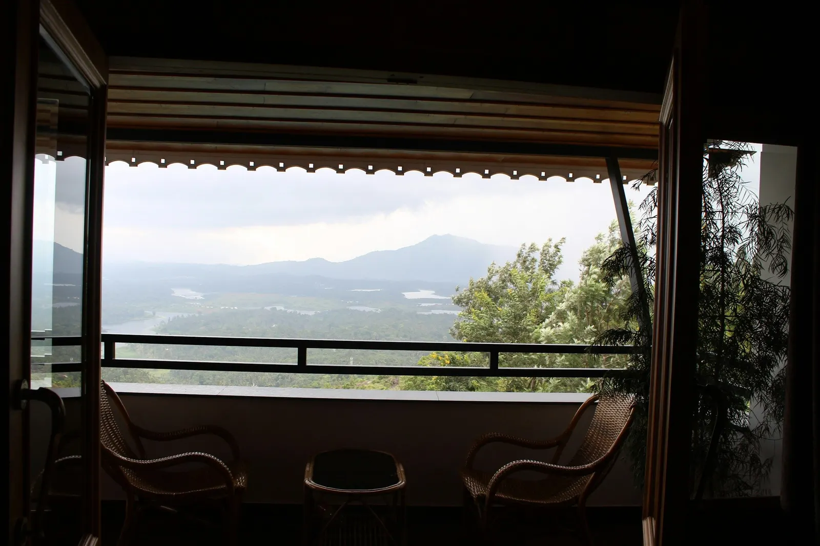Covered balcony with a bamboo chik blind rolled up under the slab, wicker chairs and a misty monsoon view
