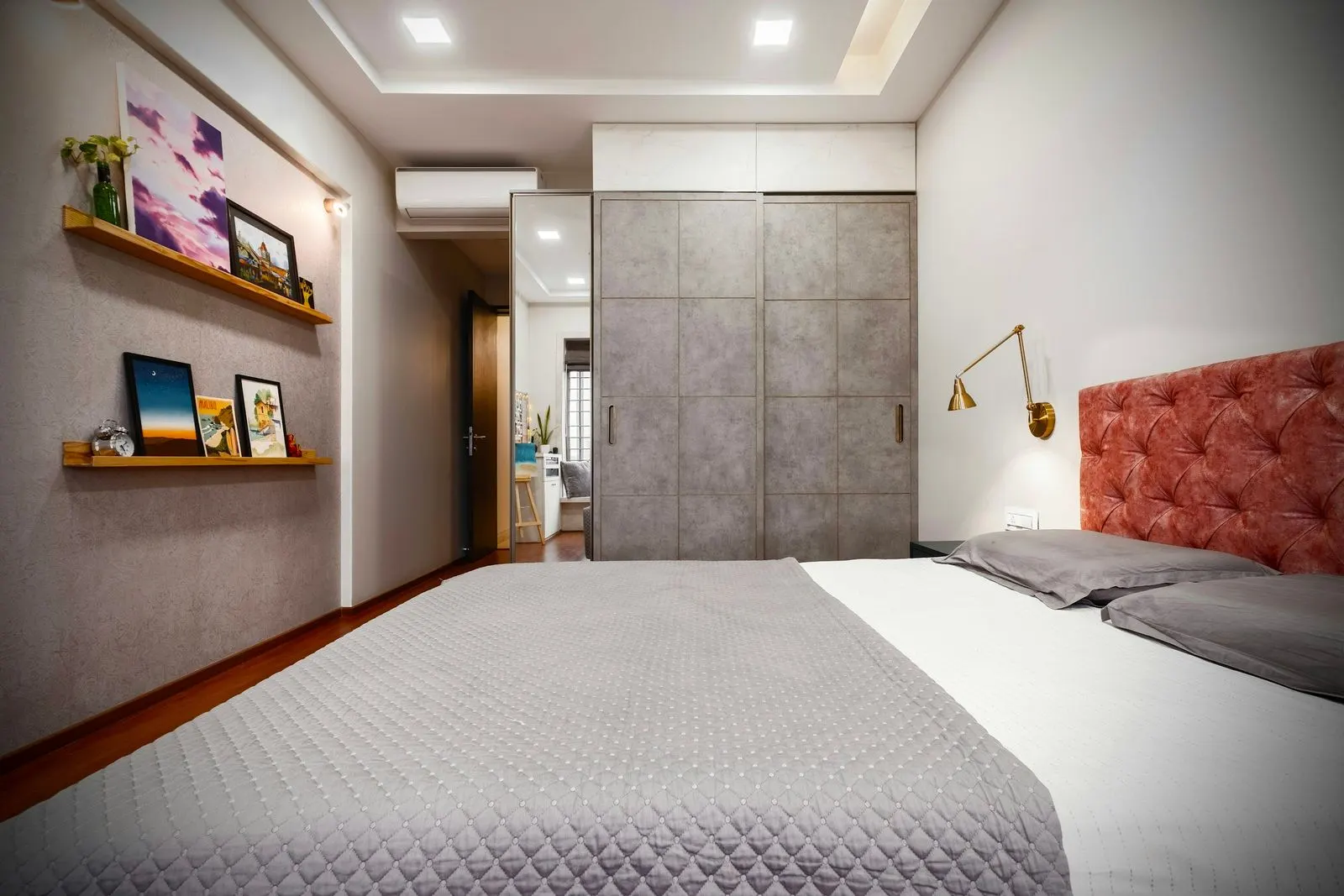 Compact bedroom with a grey full-height sliding wardrobe with one mirrored door, a split AC above the doorway, floating picture shelves and a tufted coral headboard with a brass wall lamp