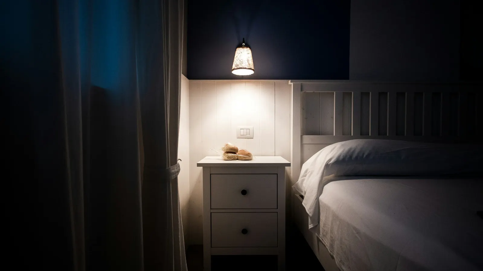 Dark bedroom lit only by a bedside lamp, with a heavy curtain drawn across the window