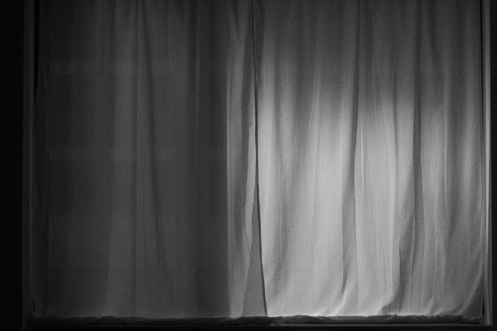 Closed white curtains in a dark room with daylight glowing through the fabric and along the bottom edge