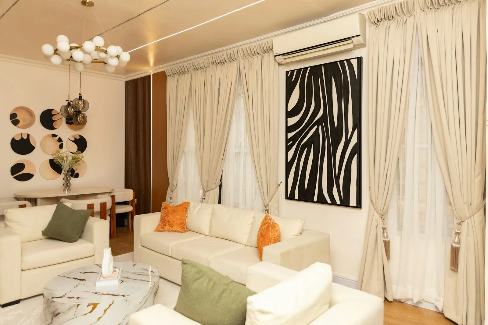 Living room with floor-length beige pencil-pleat curtains held back with tassel tiebacks over white sheers, beside a cream sofa and a large black and white artwork