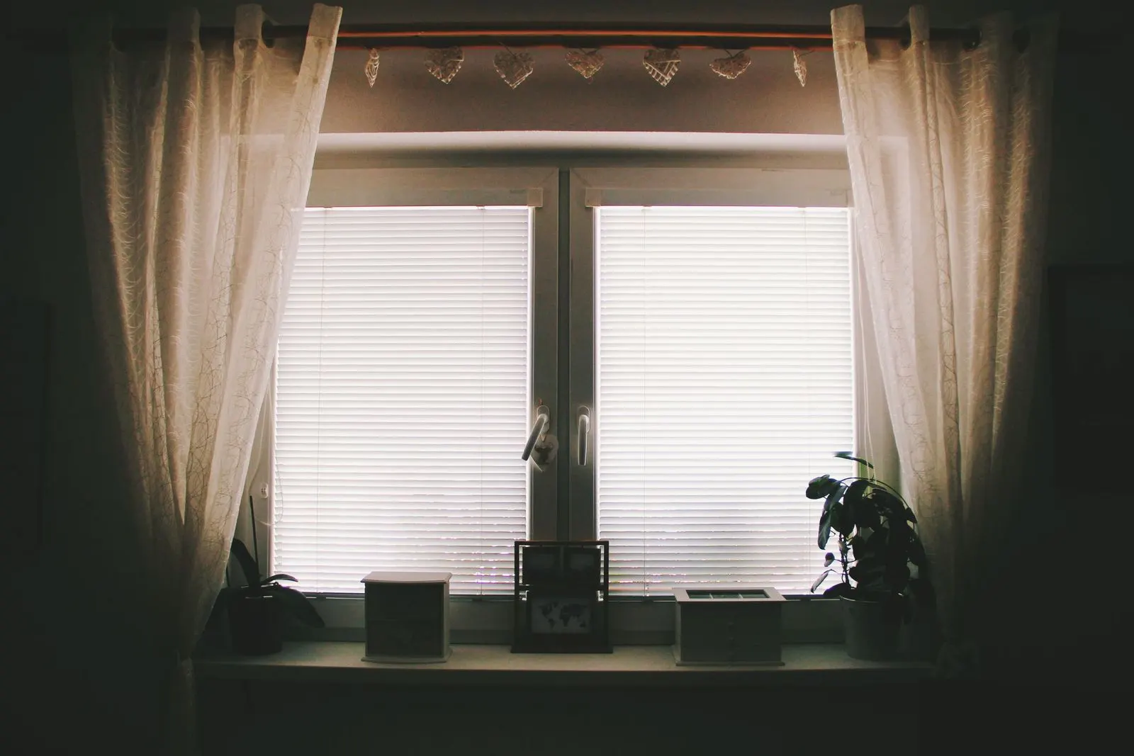 Two-pane window with white venetian blinds lowered close to the glass, framed by sheer curtains drawn to each side under a lace valance