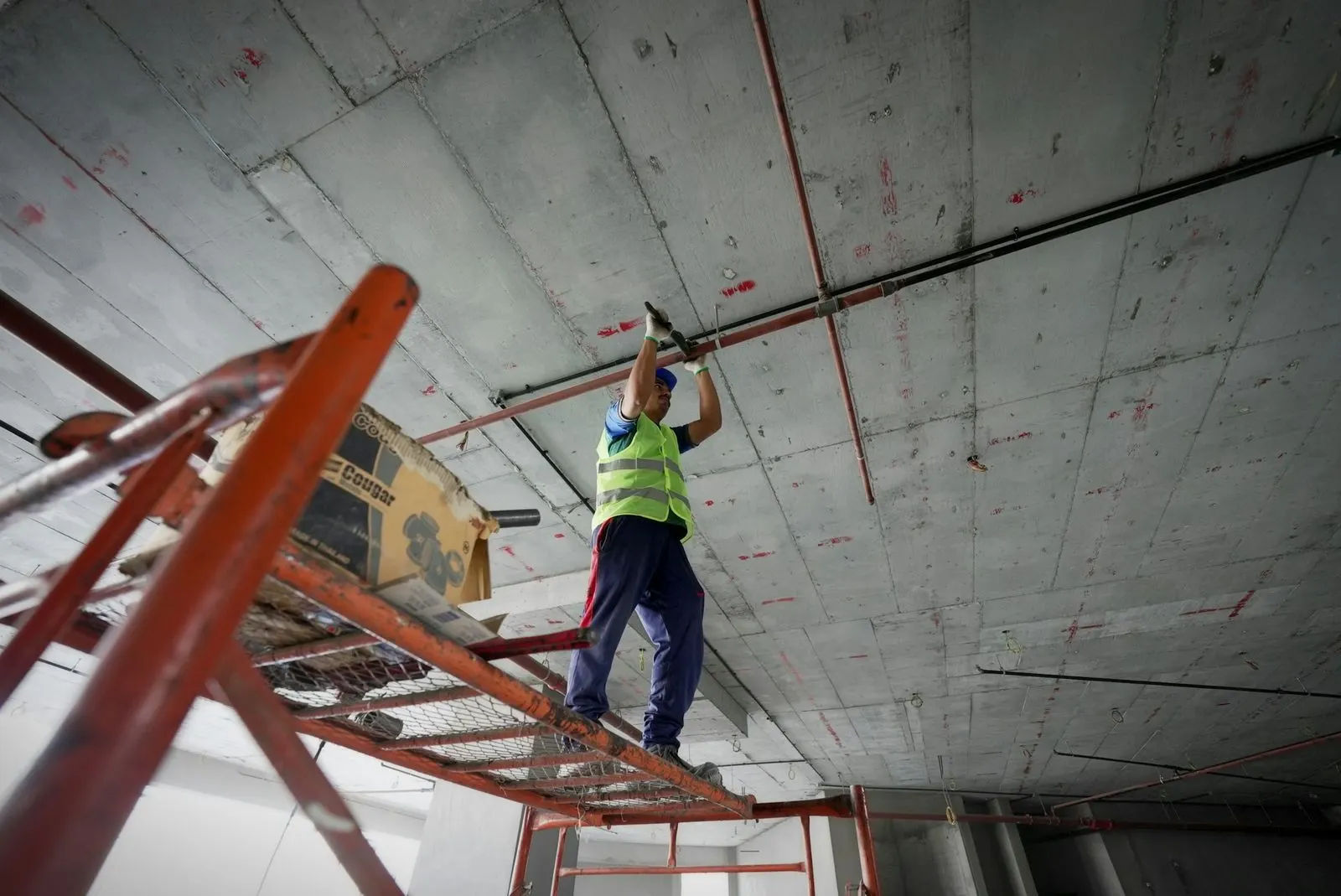 Worker on a scaffold reaching up to a bare concrete ceiling slab with exposed conduit pipes