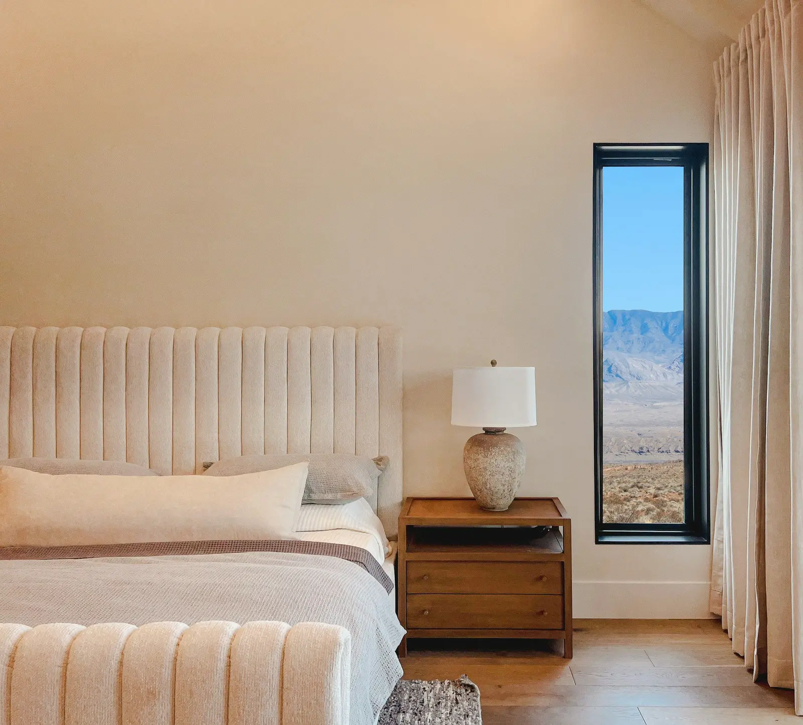 Cream vertical channel-tufted headboard behind a made bed, with a wooden bedside table and lamp