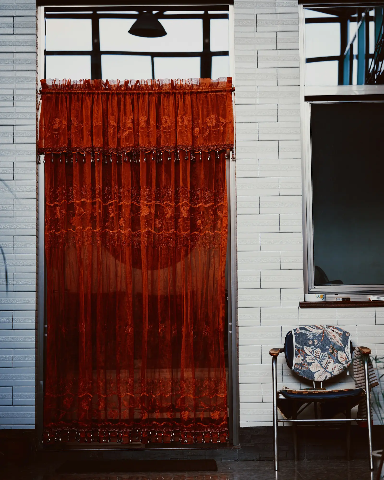 Rust-red patterned sheer curtain with a beaded valance hung across a doorway beside a white tiled wall