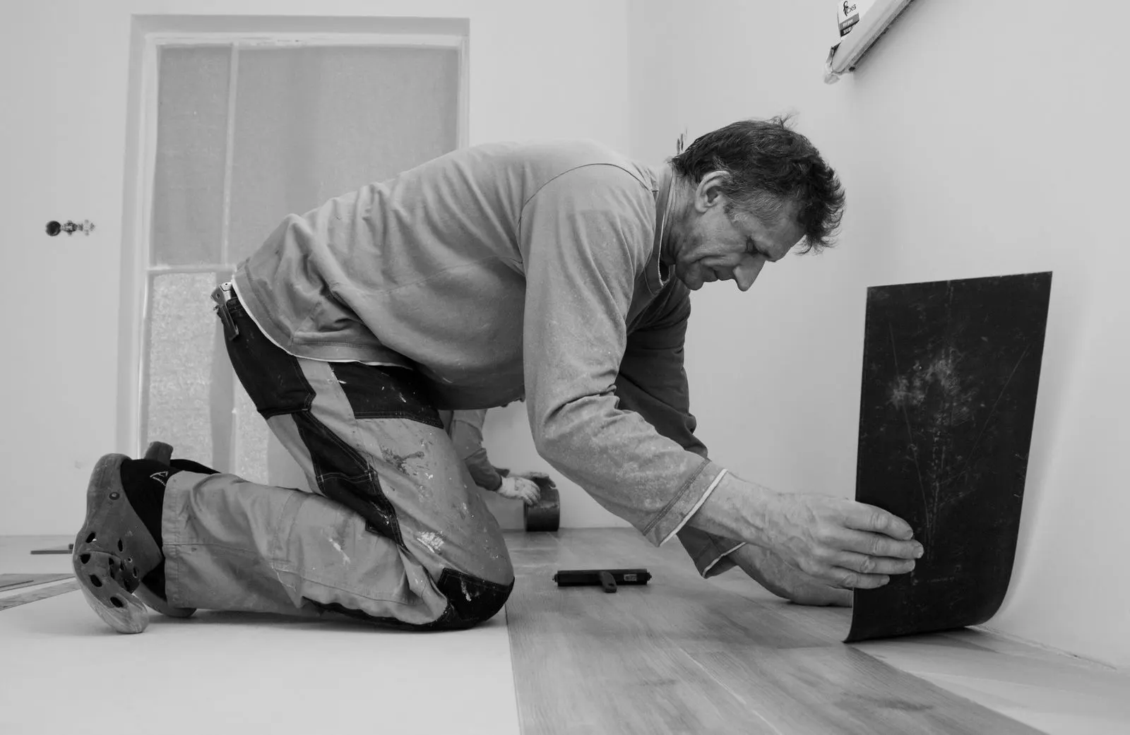 Black and white photo of a flooring fitter kneeling to lay a vinyl plank against the wall, with laid planks behind him