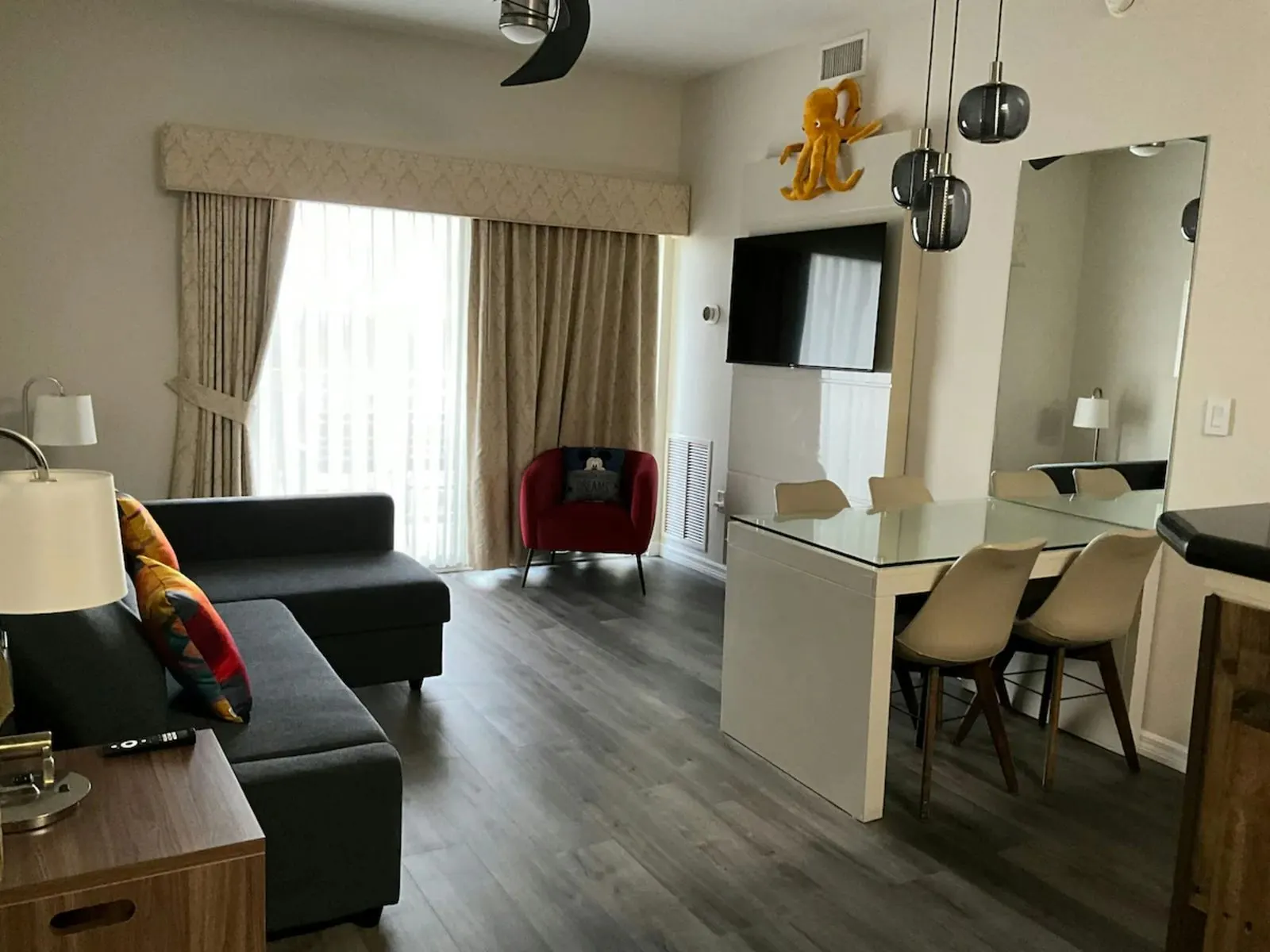 Open-plan living and dining area with curtains under a fabric pelmet at a glass door, a corner sofa, a wall-mounted TV and a glass dining table