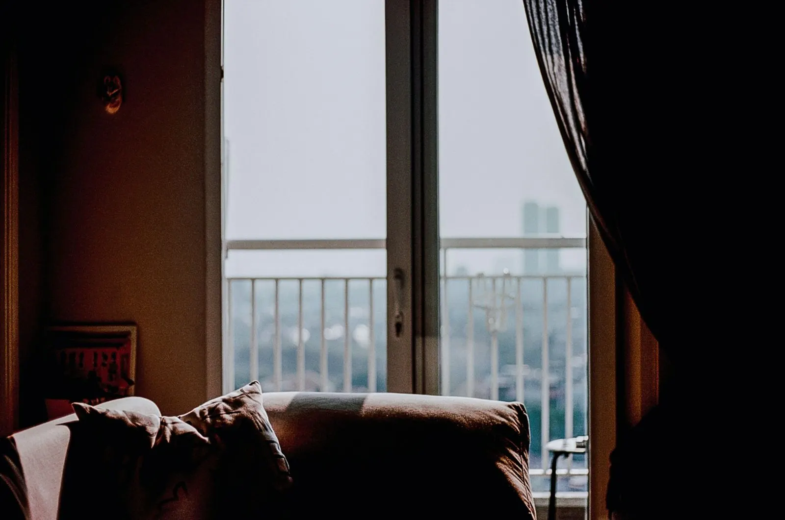 Sofa beside a full-height glass door with a balcony railing and distant towers in the haze beyond