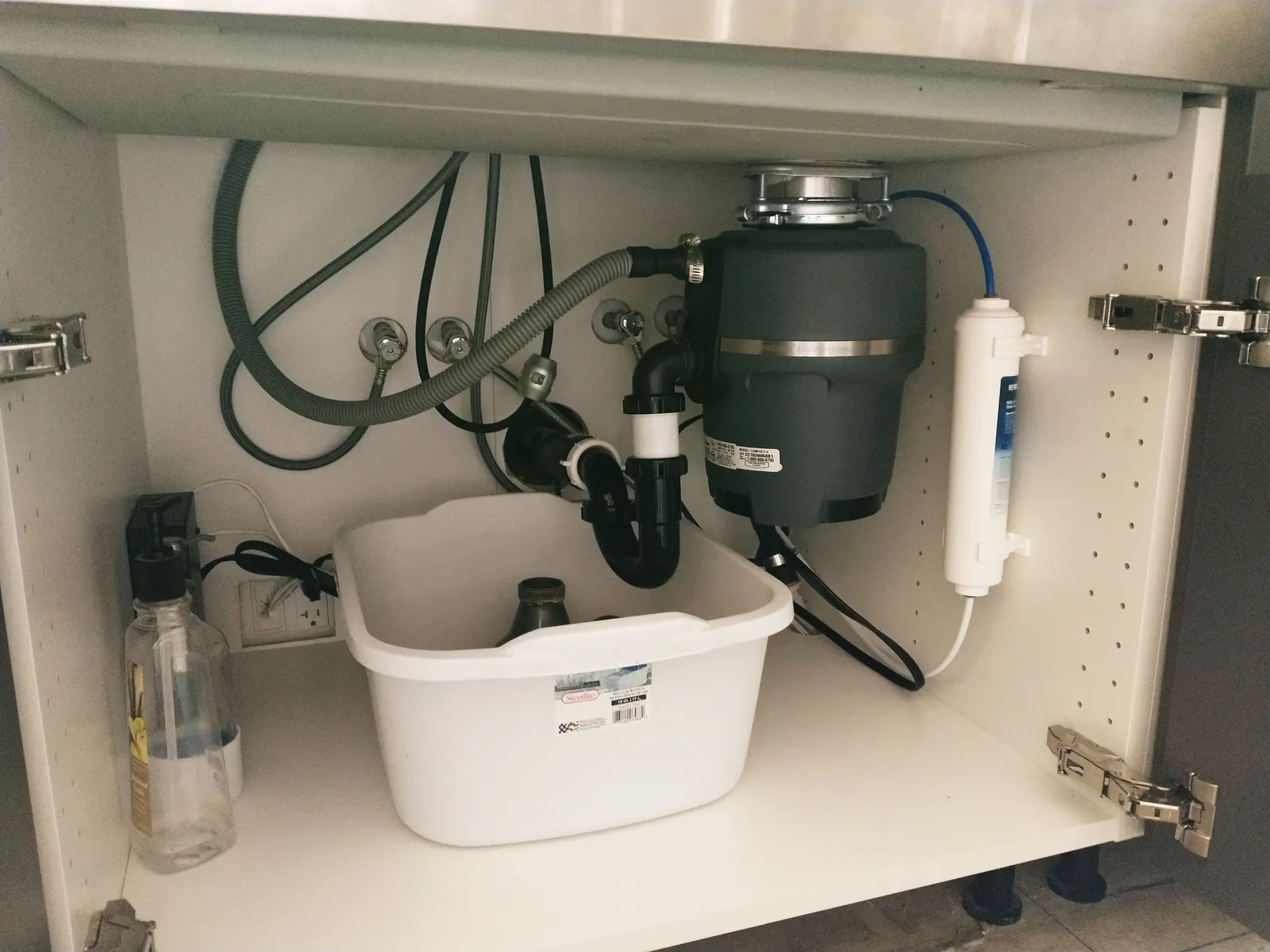 Open sink base cabinet showing the waste pipe, disposal unit, water filter and soft-close hinges inside a white carcass