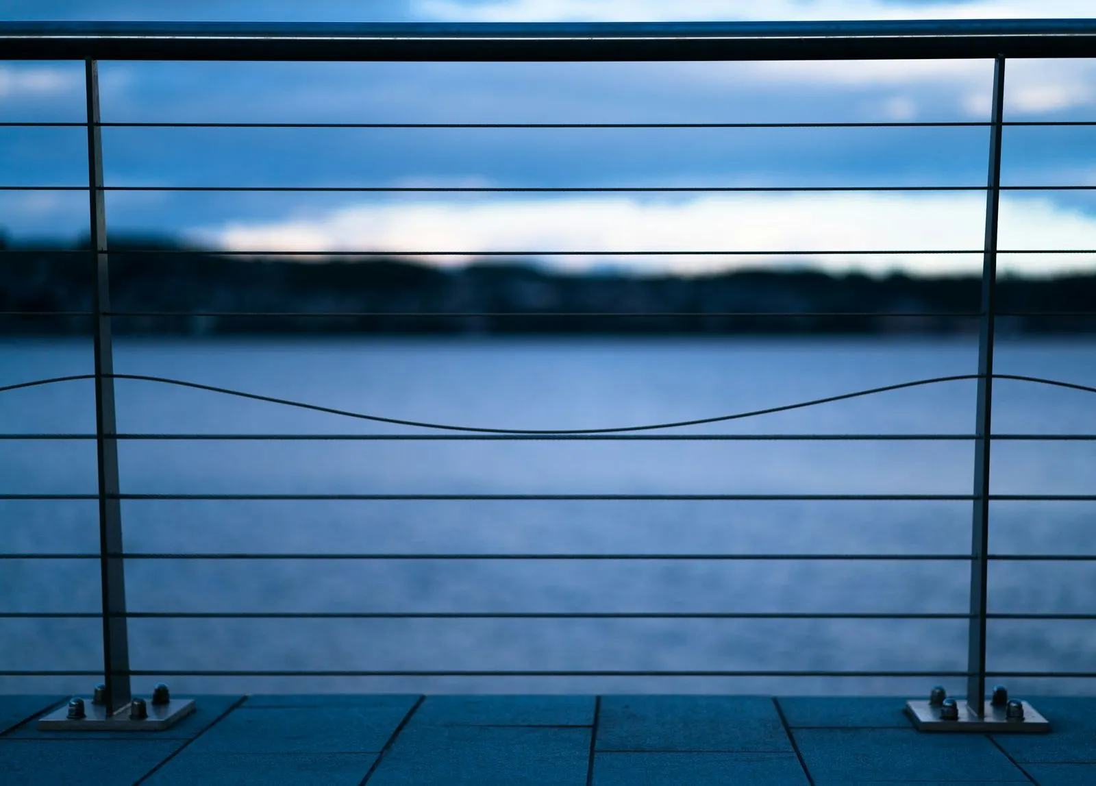 Horizontal stainless steel cable railing on a waterfront balcony at dusk, with the water and far shore blurred behind