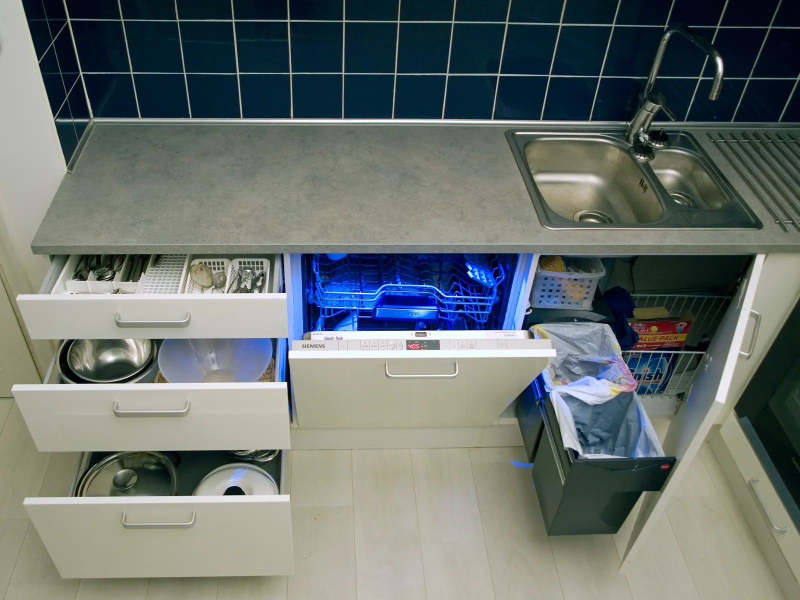Base units under a stainless steel sink pulled open: drawers of pans and pots, an integrated dishwasher and an under-sink cabinet with pull-out waste bins