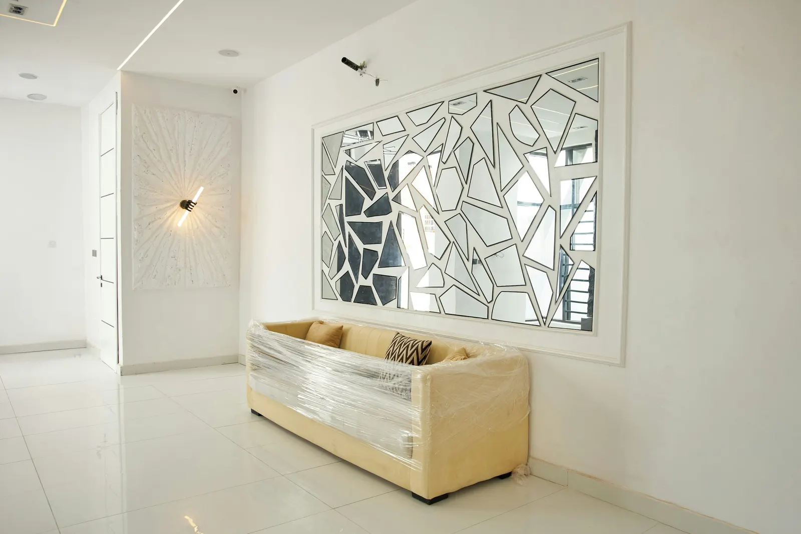 Newly delivered beige sofa still wrapped in protective plastic film in an apartment with white floor tiles and a mirrored wall panel