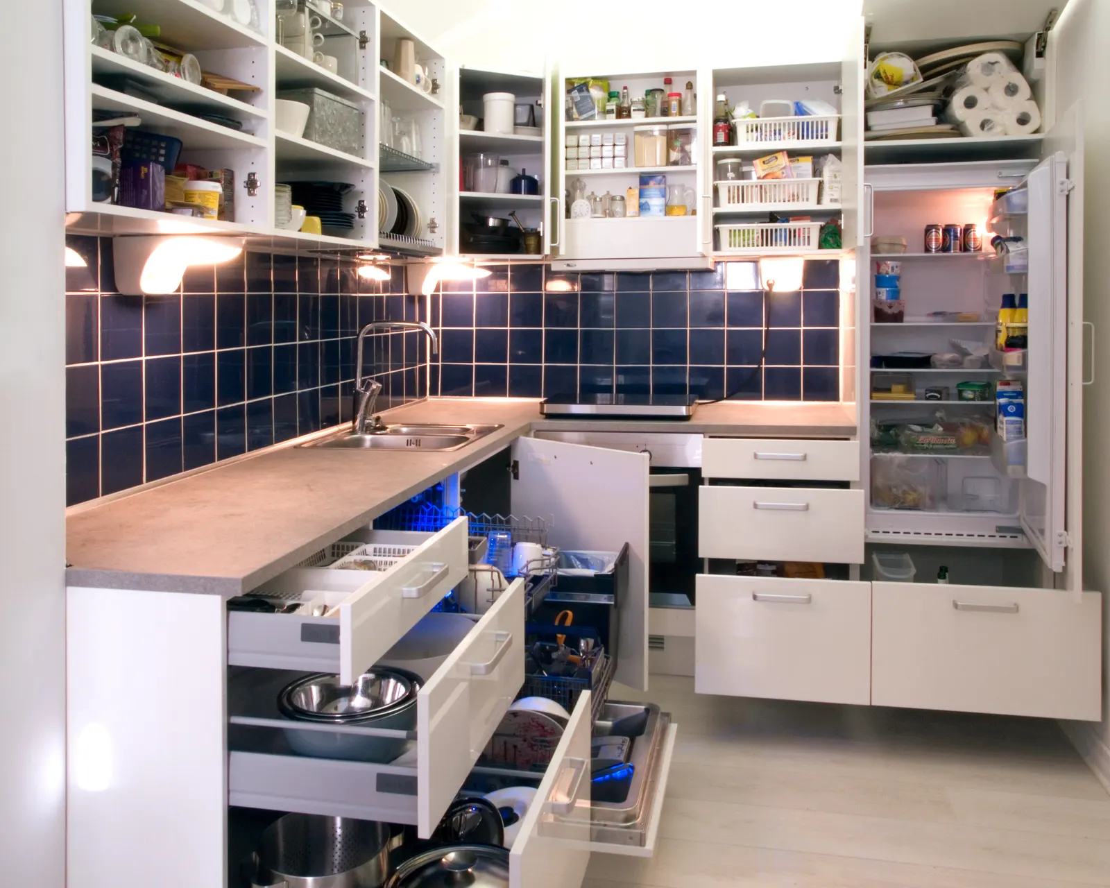 Compact white modular kitchen with every drawer and door open: deep base drawers hold steel bowls, pots and lids, next to an open dishwasher and an integrated fridge