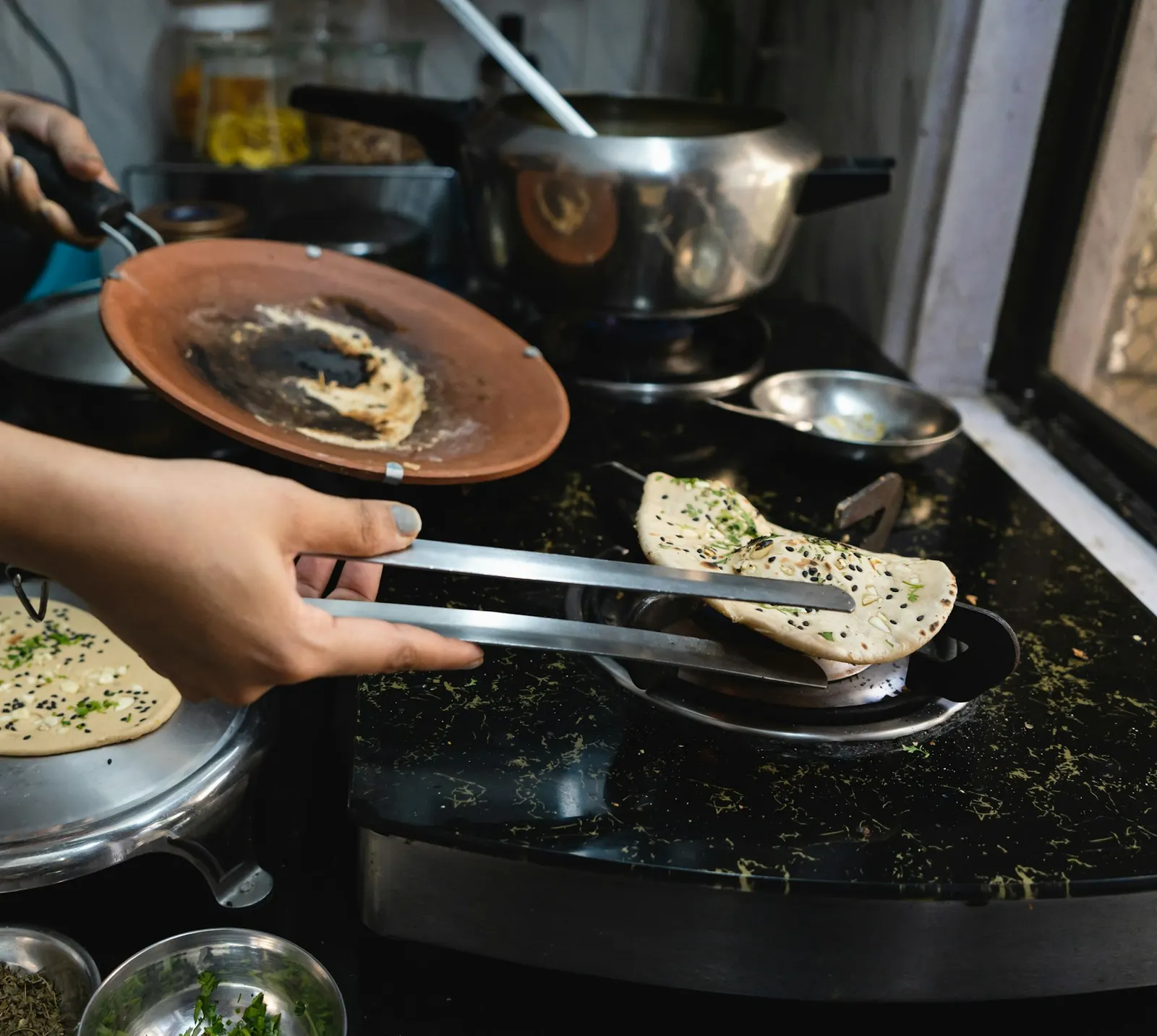 Hands turning a paratha with steel tongs on a black stone counter beside the hob, with a clay tawa, a steel pressure cooker and small steel bowls around it
