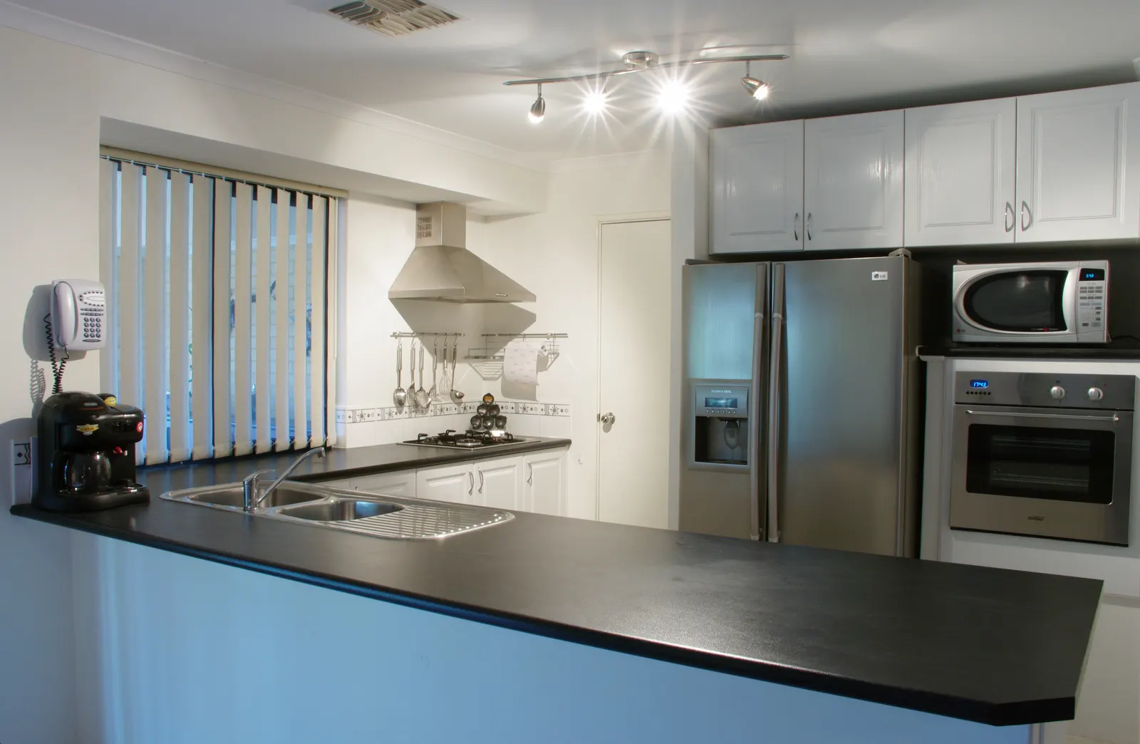 Kitchen with a double sink set into a long front counter facing a back wall with a double-door fridge, a microwave and a built-in oven, and a gas hob under a steel chimney on the side wall