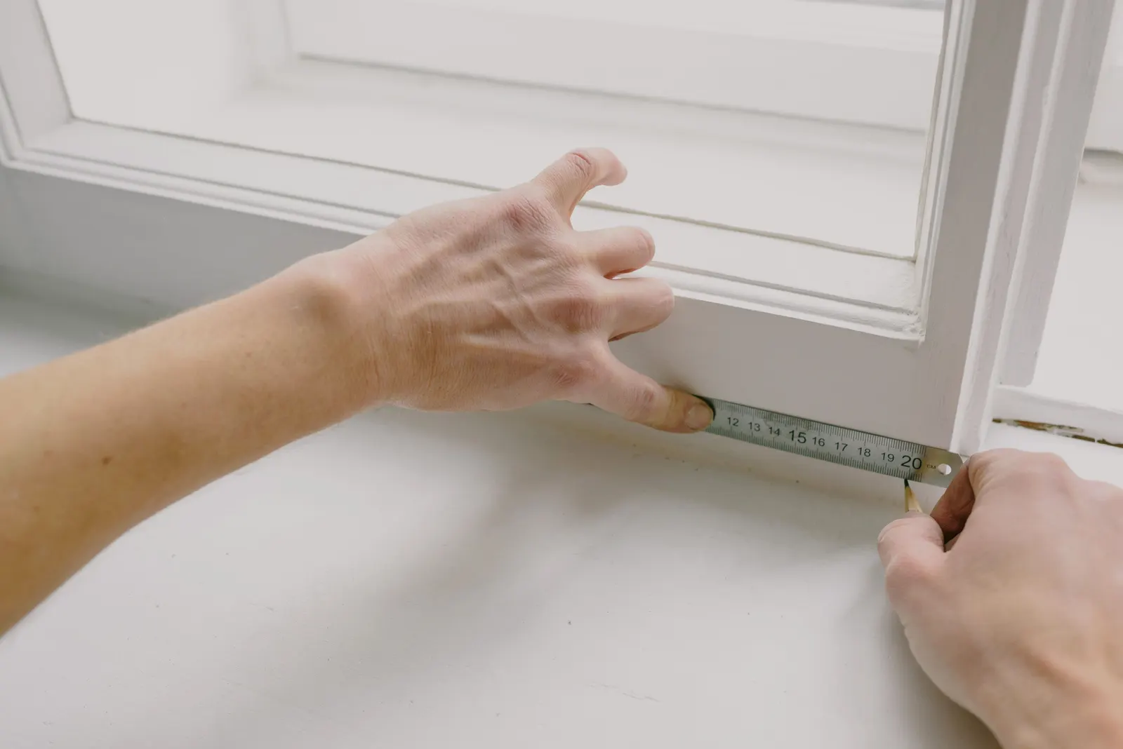 Hands measuring the inside of a white window frame with a steel rule and marking it with a pencil