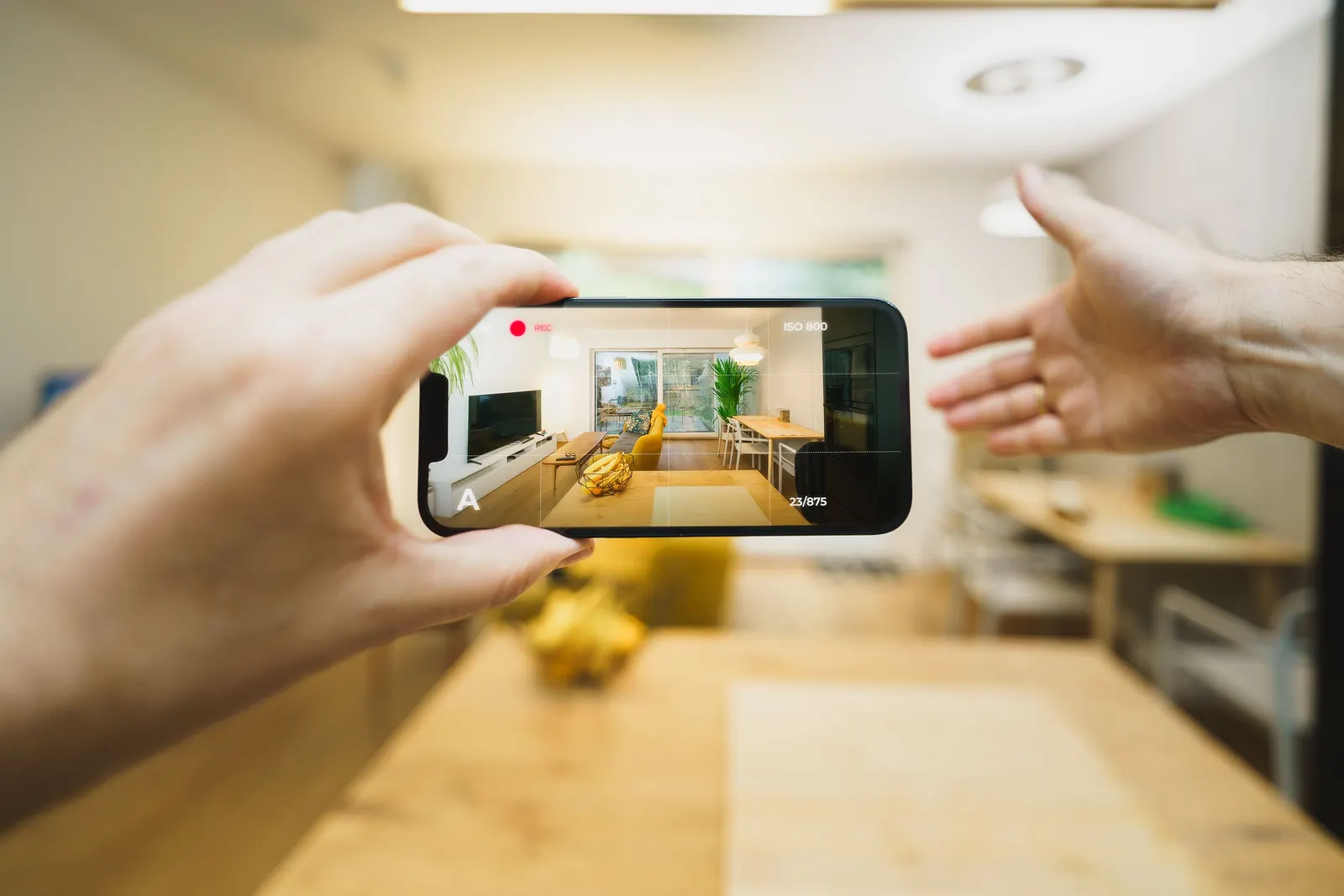 Hand holding up a phone that is filming a walk-through of a bright living room and kitchen