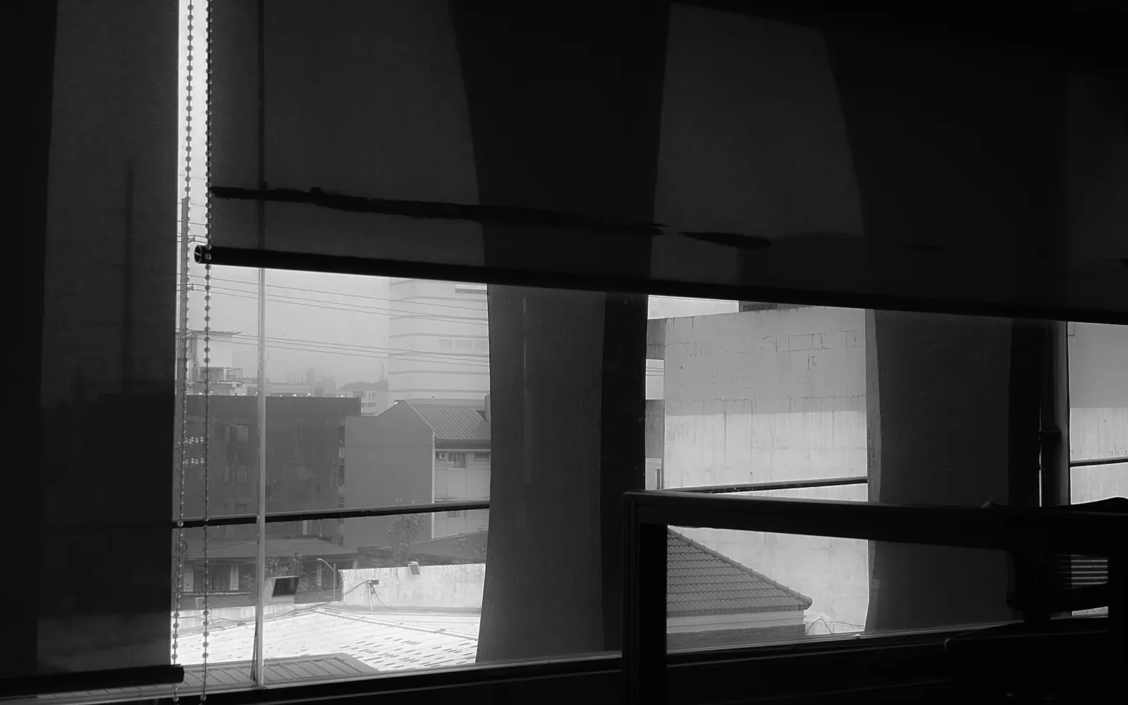 Sunscreen roller blind lowered halfway over an office window, with its chain control at the side and city buildings visible below; the outline of the columns outside shows faintly through the mesh