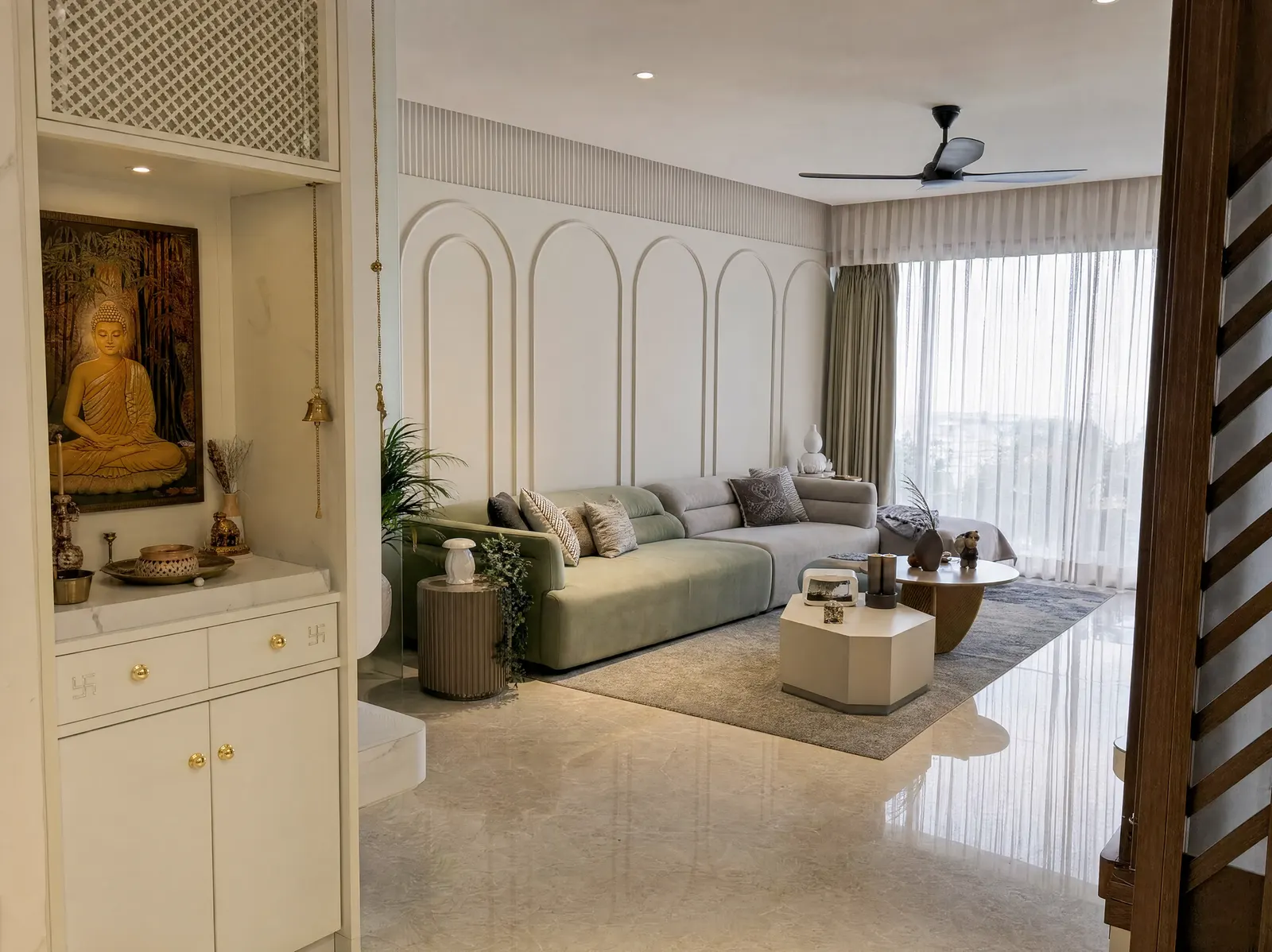 Built-in pooja niche beside a living room, with a lattice jaali panel above, a marble shelf holding brass lamps and a hanging bell, and drawers and cupboards below