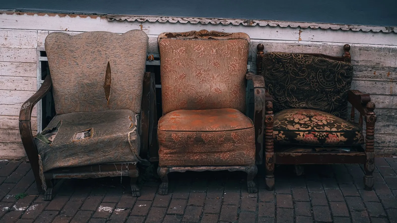 Three old armchairs with faded, torn upholstery and slumped seat cushions standing against a weathered wooden wall