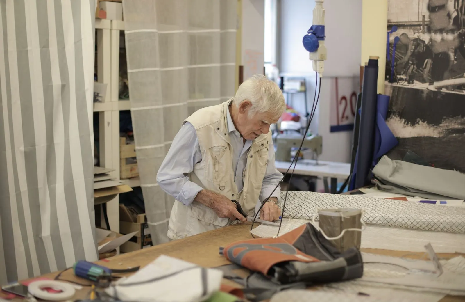Elderly craftsman cutting fabric on a large workbench in an upholstery and soft furnishing workshop