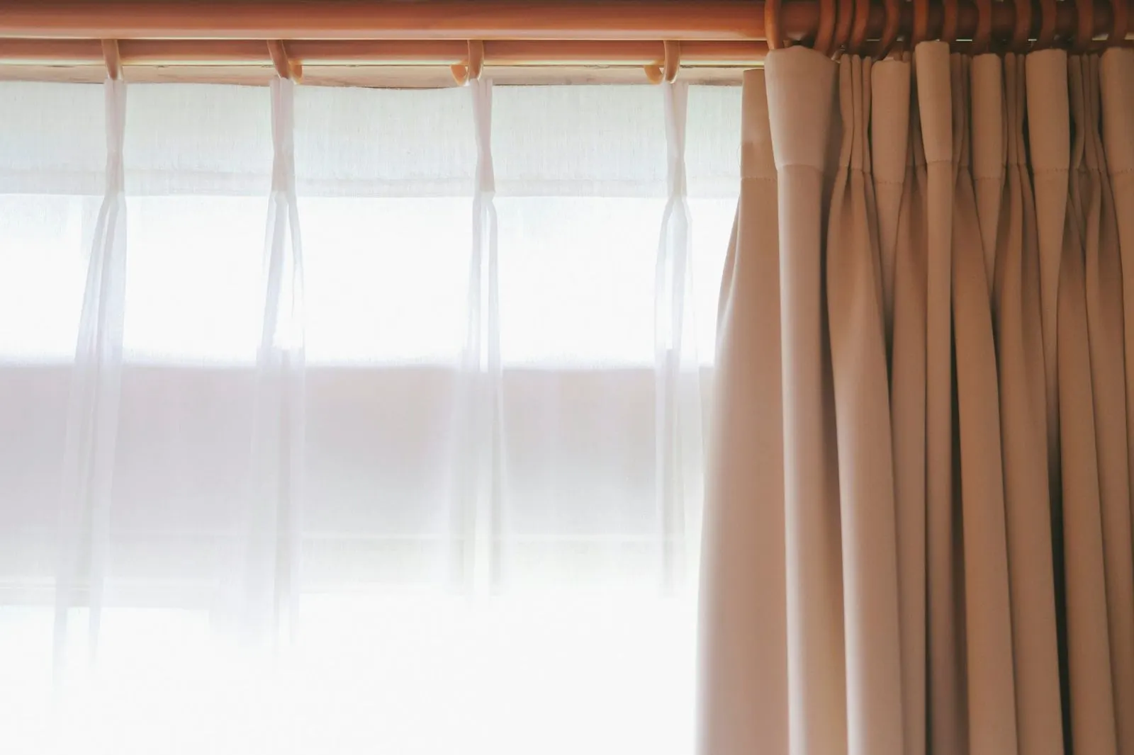 White sheer and a beige pinch-pleat curtain hanging from rings on a rod in front of a bright window