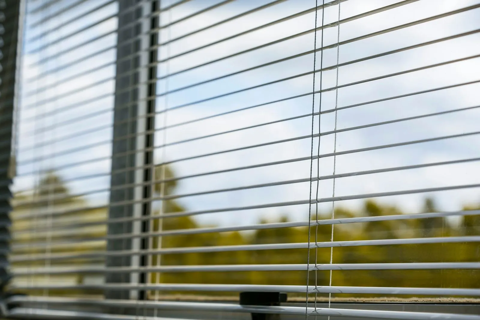 White aluminium venetian blind with its slats tilted partly open, daylight and trees showing between the slats