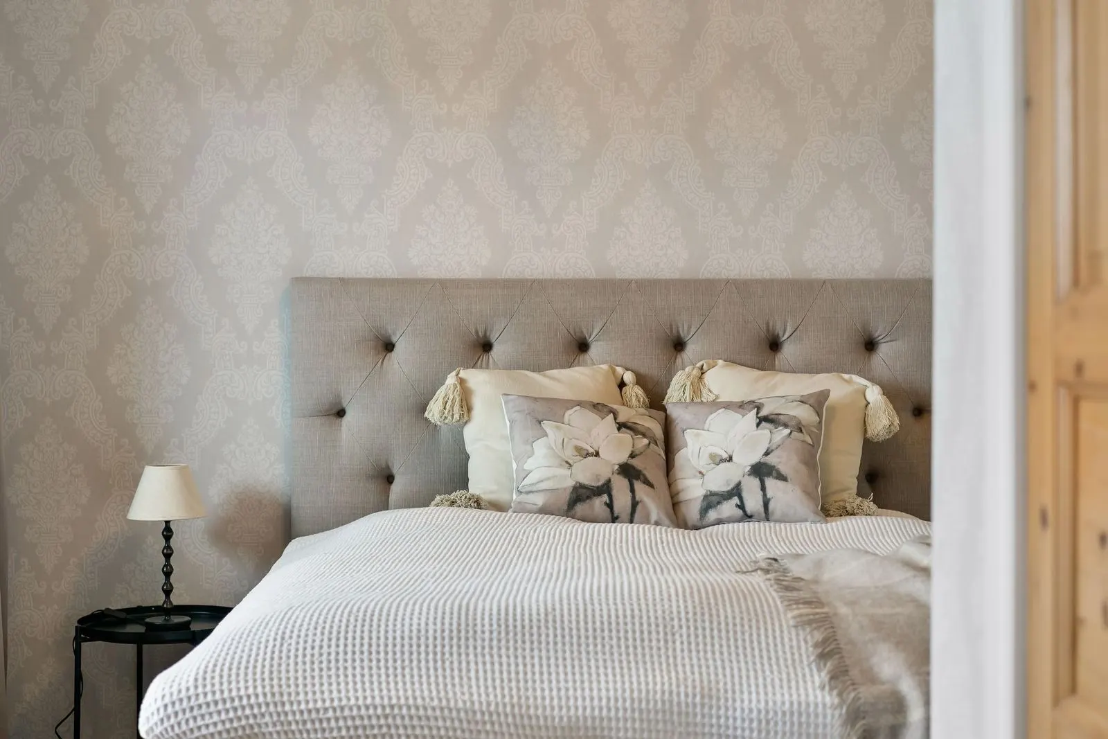 Bedroom with beige damask-pattern wallpaper behind a grey tufted upholstered headboard, floral cushions and a small bedside lamp