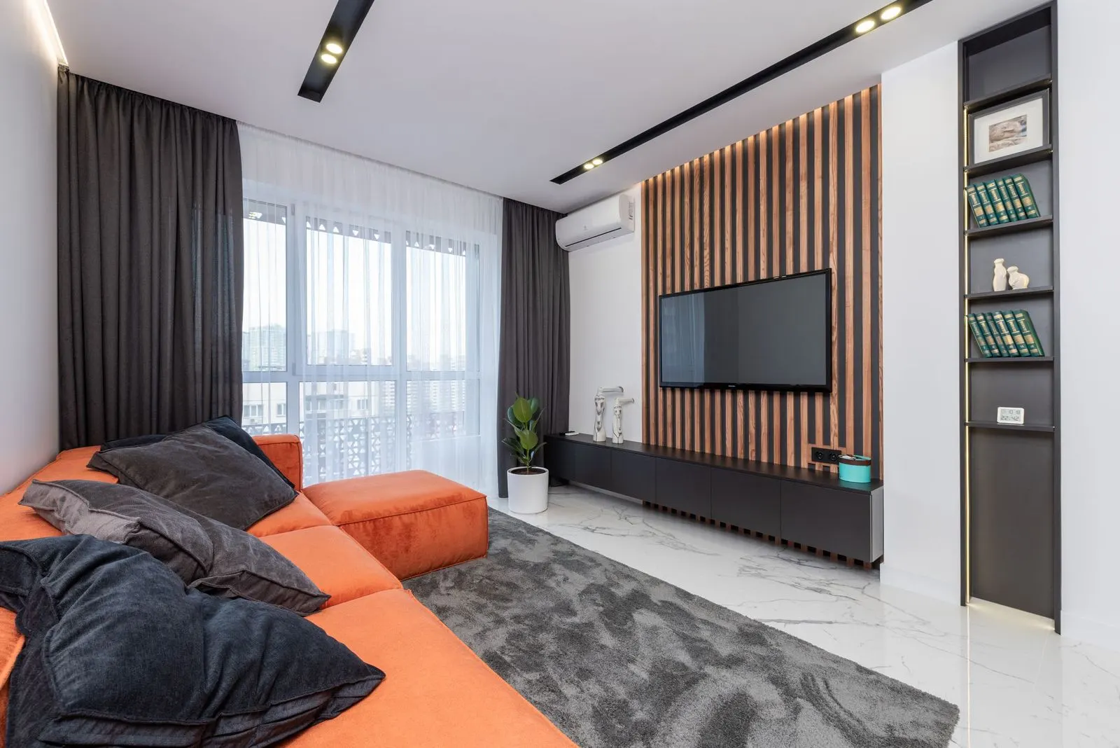 Apartment living room with a TV mounted on a wood slat feature wall above a low black unit, an orange sectional sofa, grey curtains over sheers and a high-rise view