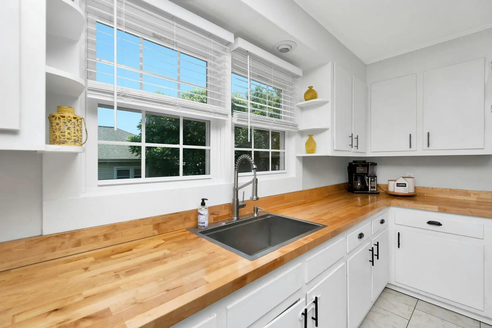 White venetian blinds raised partway over two kitchen windows above a steel sink and a wooden worktop