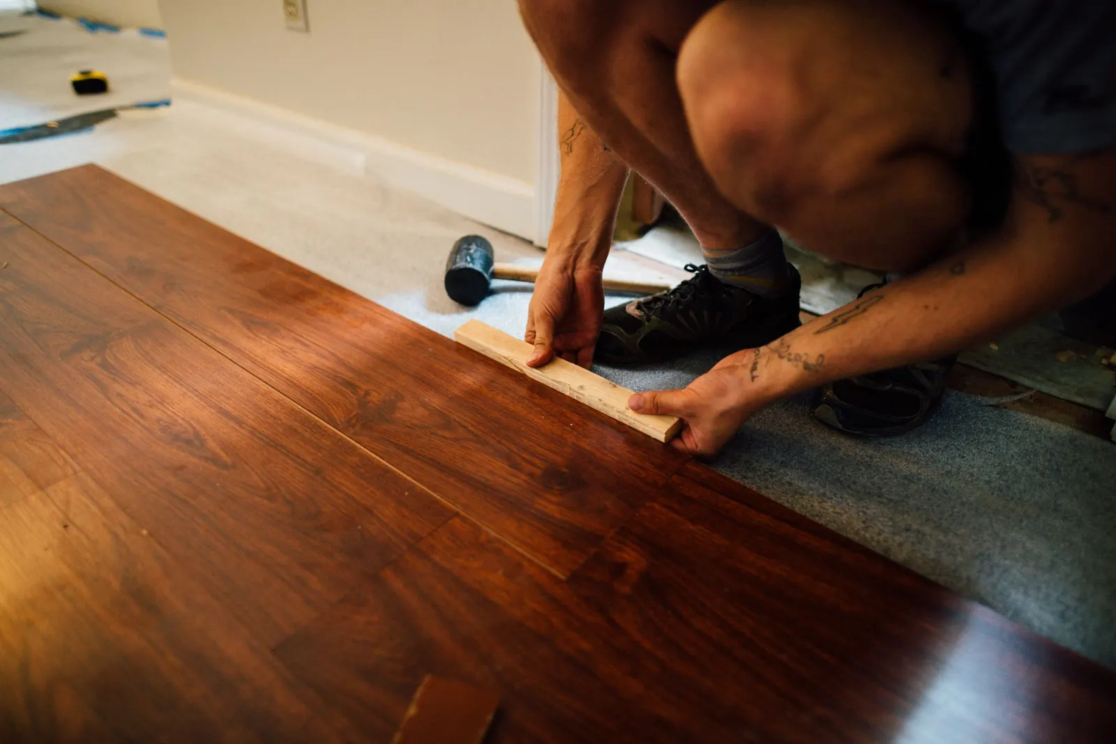 Installer tapping a dark wood plank into place with a tapping block, a mallet and underlay beside him