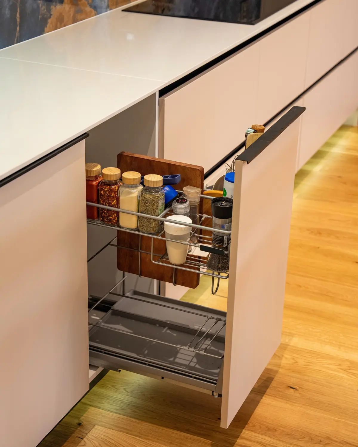 Narrow pull-out beside an induction hob holding spice jars, a pepper grinder and a chopping board in a wire frame