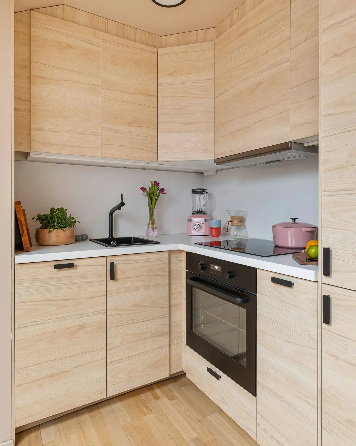 Compact L-shaped kitchen with light wood-grain shutters on the wall and base units, black bar handles and a white counter