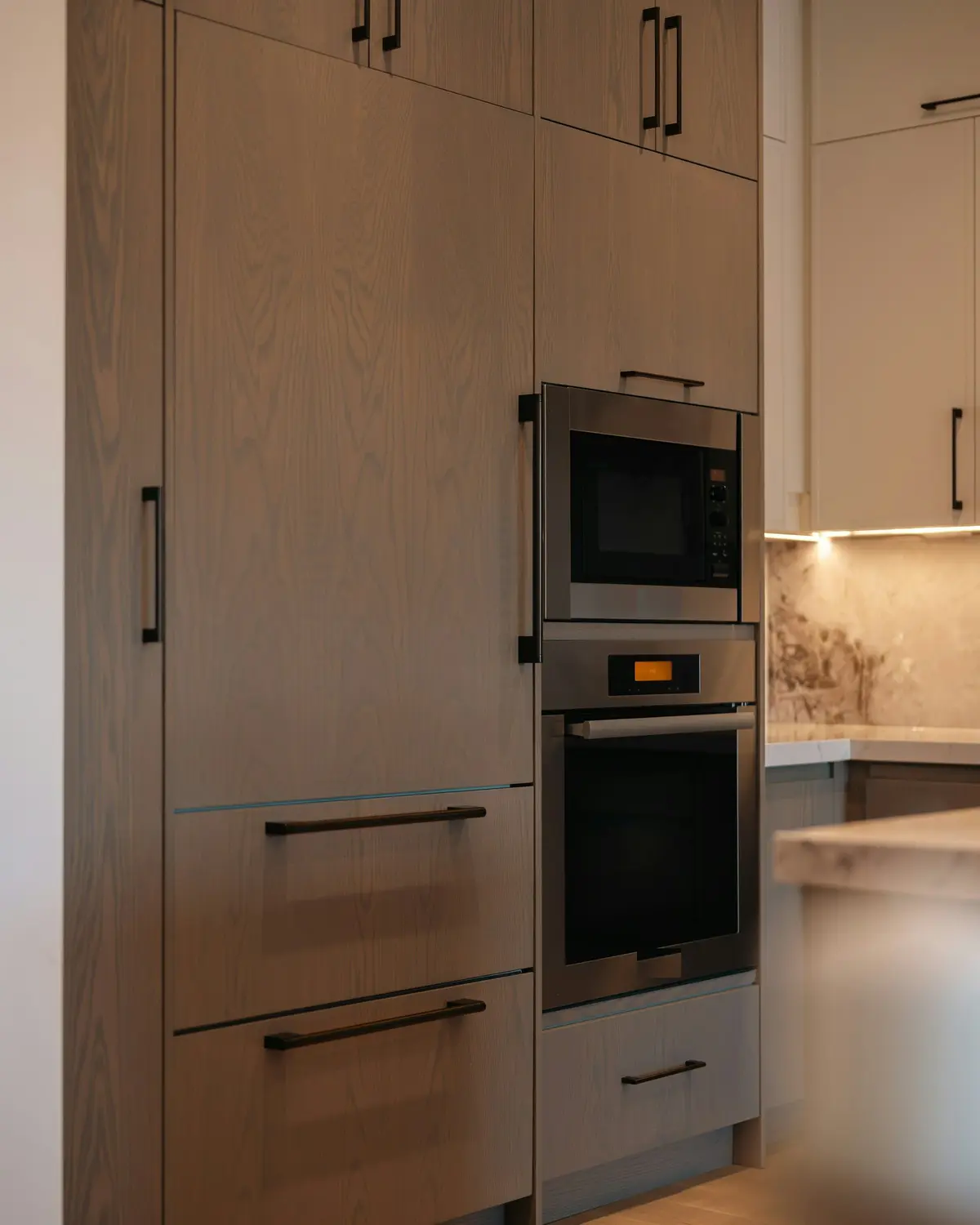 Built-in microwave above a built-in oven in a tall wood-finish unit, with lights under the wall cabinets beside it