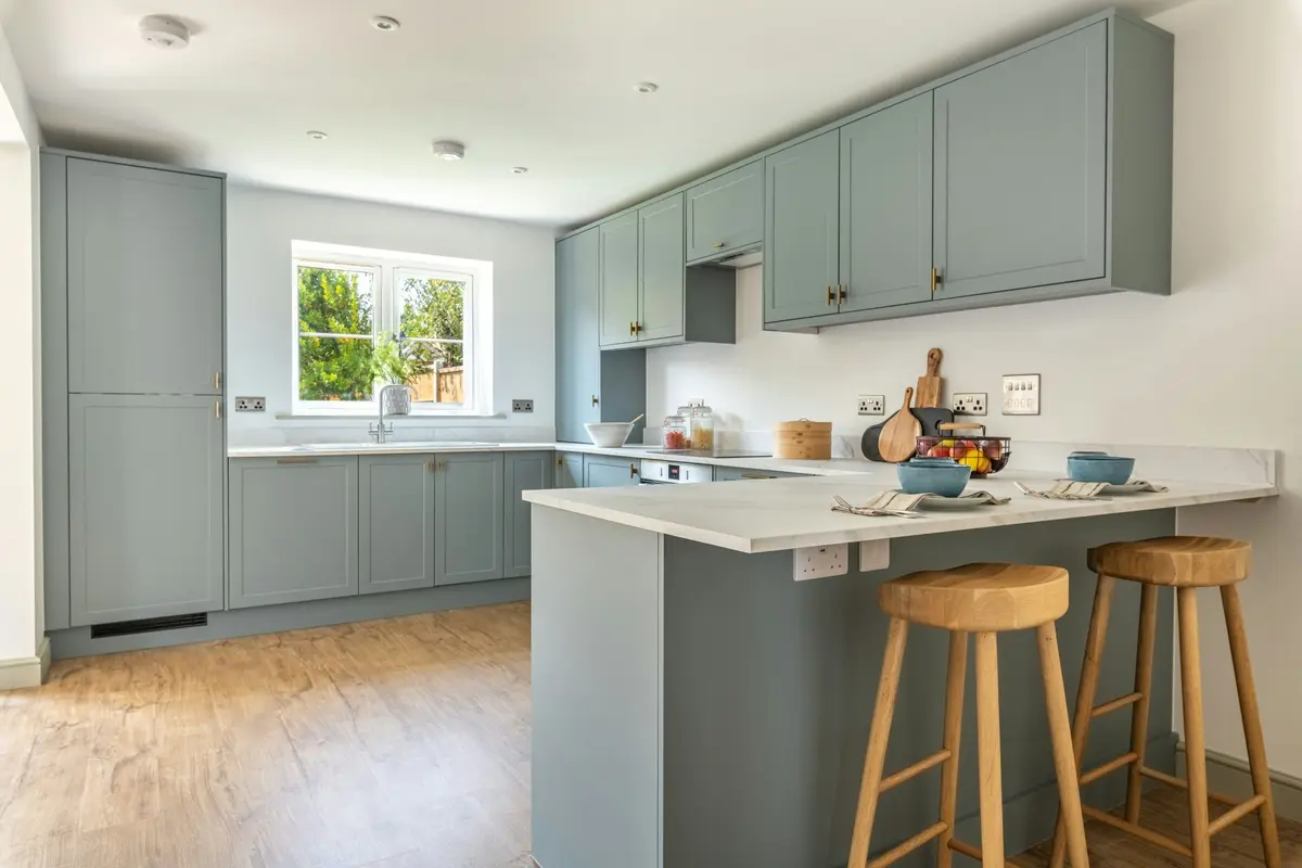 Sage green L-shaped kitchen with a breakfast counter that juts out from one run, with two wooden bar stools