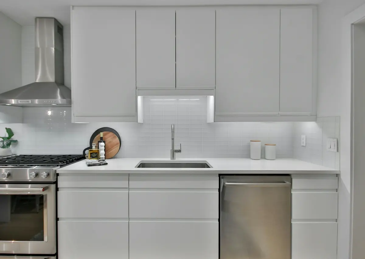 Single-wall kitchen with white handle-less cabinets, a range and hood at one end, an undermount sink and a dishwasher in one straight run