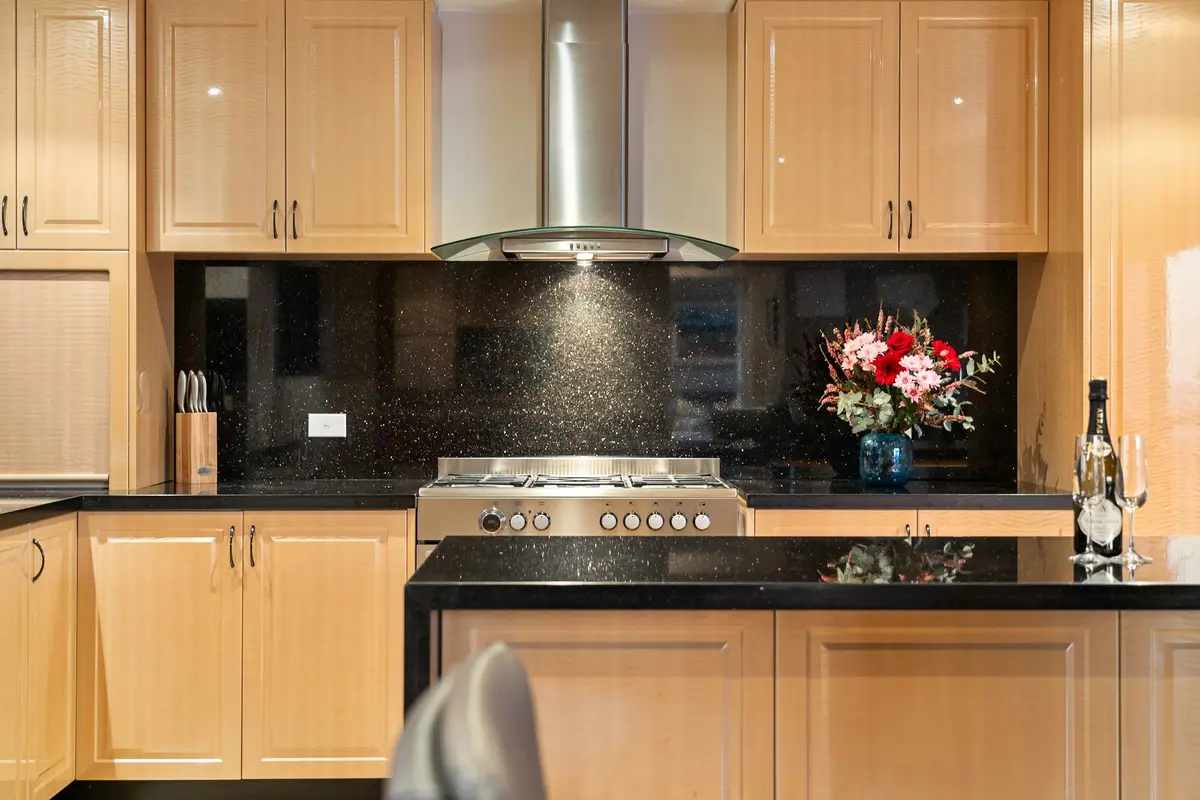 Black granite counter and full-height backsplash with gold flecks, a stainless steel range and chimney hood, and light wood cabinets
