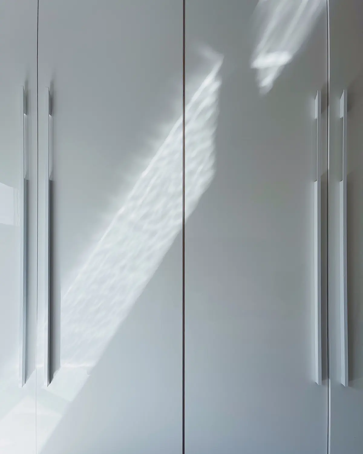 Close-up of white high-gloss wardrobe doors with long vertical handles, reflecting a band of sunlight