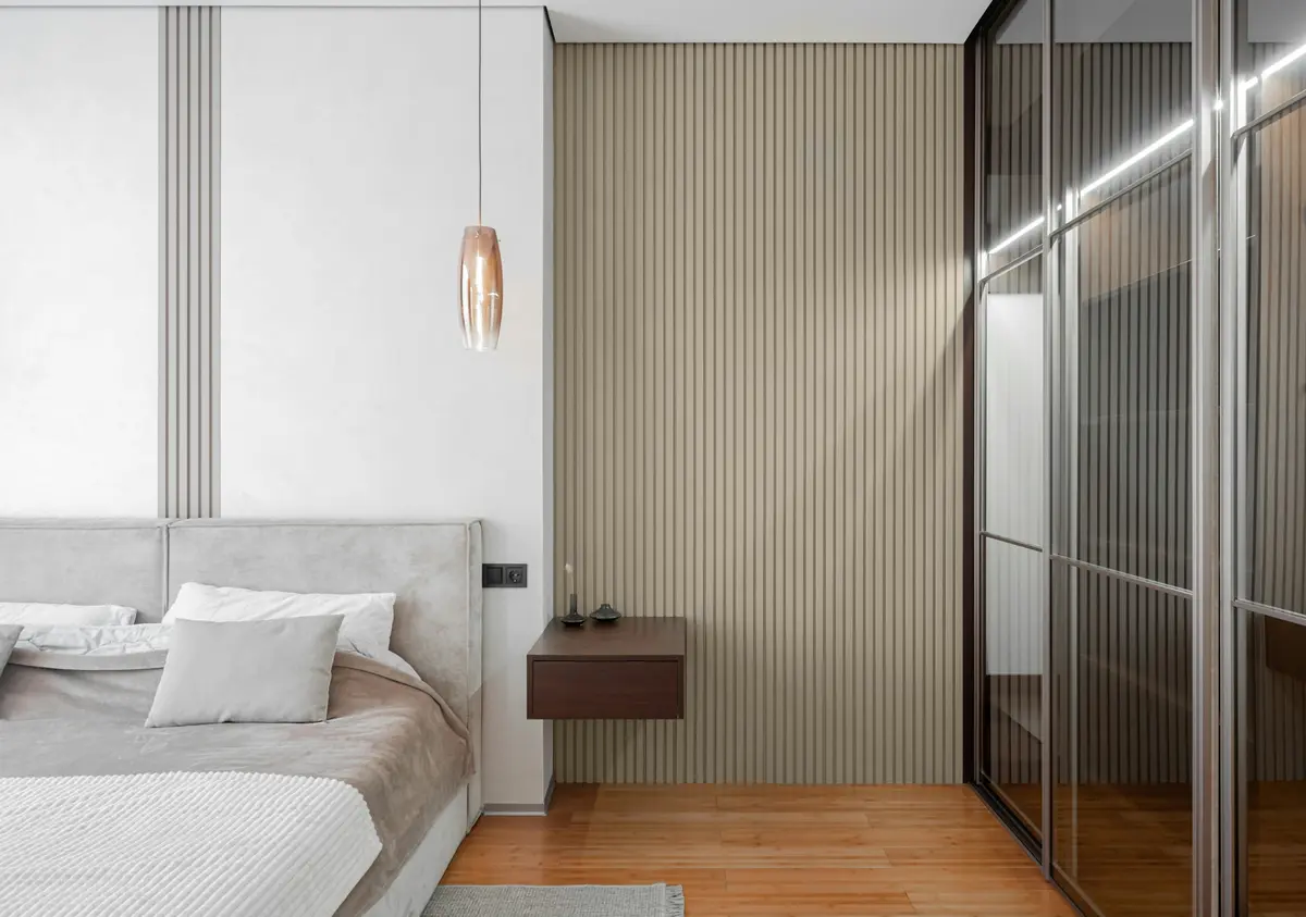 Bedroom wardrobe with tinted glass doors in slim dark aluminium frames, lit from inside, beside a slatted wall