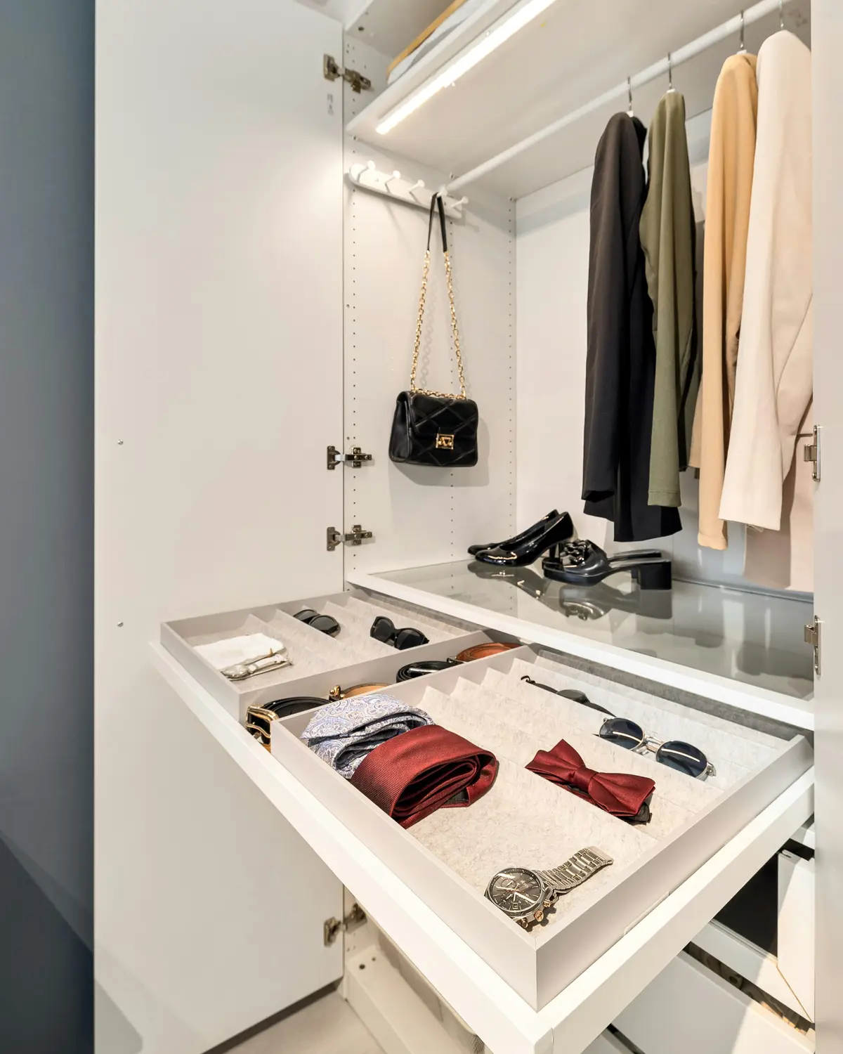 Open wardrobe with a pull-out accessory tray holding ties, a bow tie, sunglasses and a watch, below a hanging rail and LED light