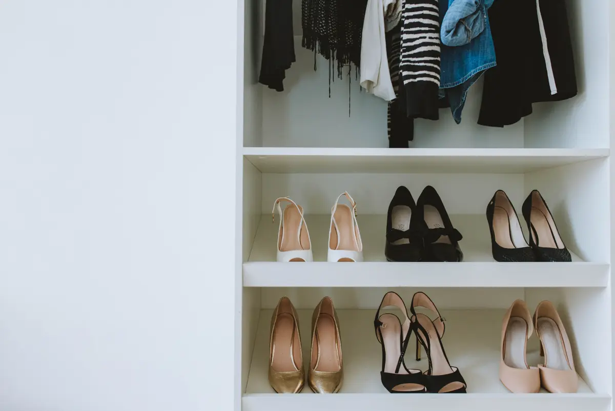 Two white shelves of heels inside a wardrobe, below a rail of hanging clothes