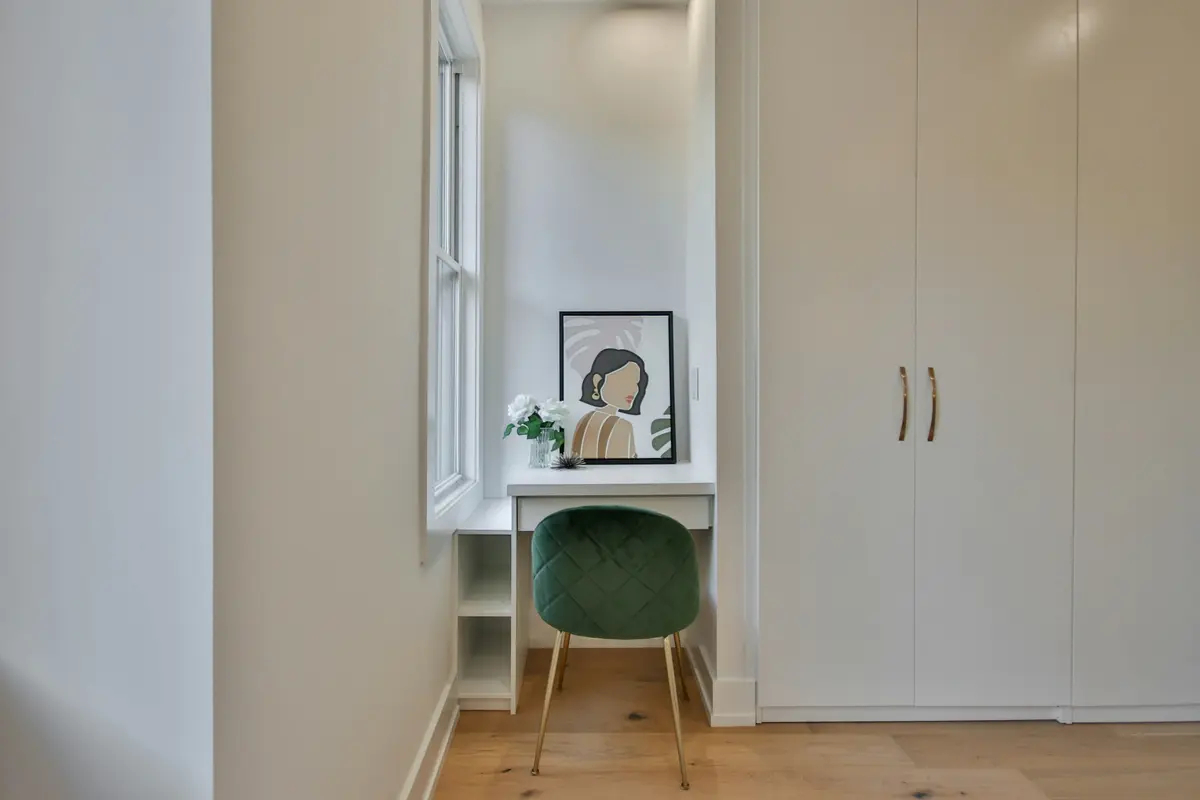 White full-height wardrobe beside a built-in desk niche with a green velvet chair under a window
