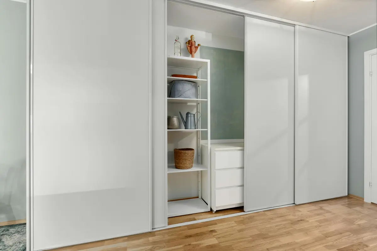 Wall-to-wall white sliding wardrobe with one panel pushed aside to show open shelves and a small chest of drawers