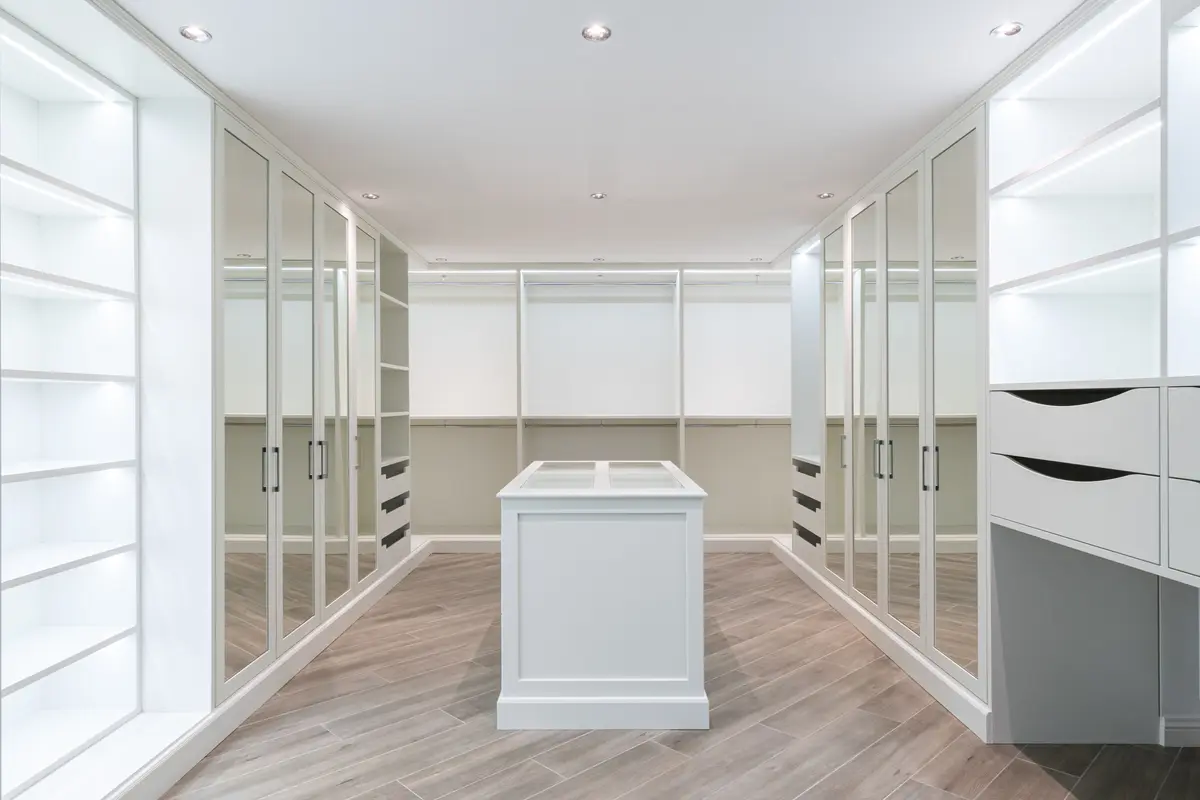 White walk-in wardrobe with lit open shelves, mirrored doors on both sides and a white island in the middle
